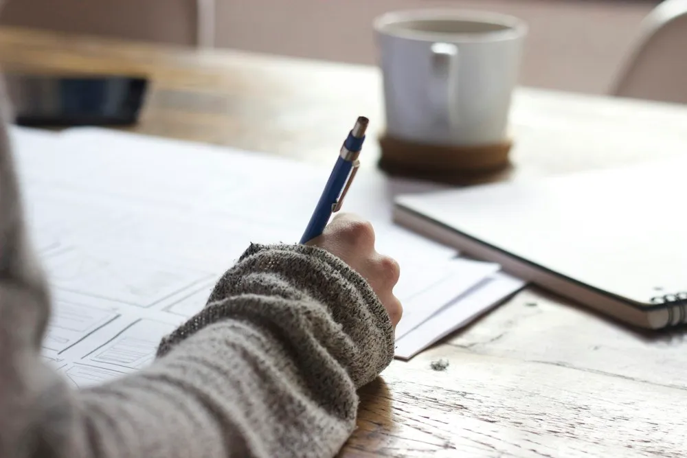 Student writing notes with a pen in a notebook beside a cup of coffee