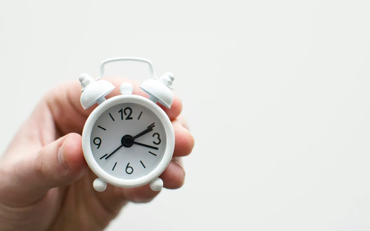 Hand holding a small white alarm clock against a plain background